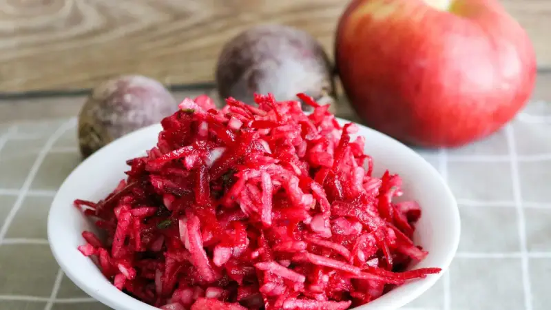 Frischer Salat aus rote Bete raspeln, mit einem Apfel und ganzen Rote Beten im Hintergrund.