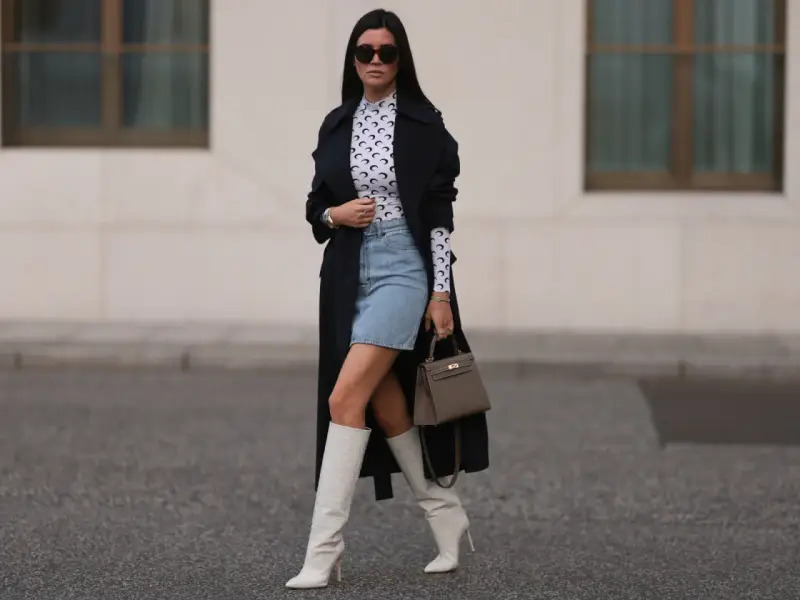 Outfit per uscire con un ragazzo: gonna di jeans, top a collo alto con stampa luna, trench nero, stivali bianchi alti e borsa grigia.