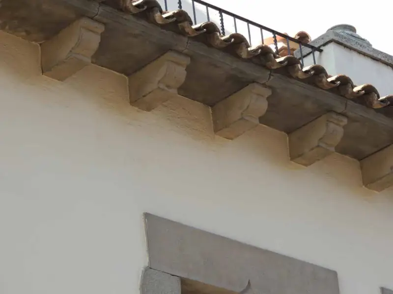 Detalle de qué es una cornisa, con ménsulas de piedra decorativas bajo tejas curvas y una barandilla metálica.