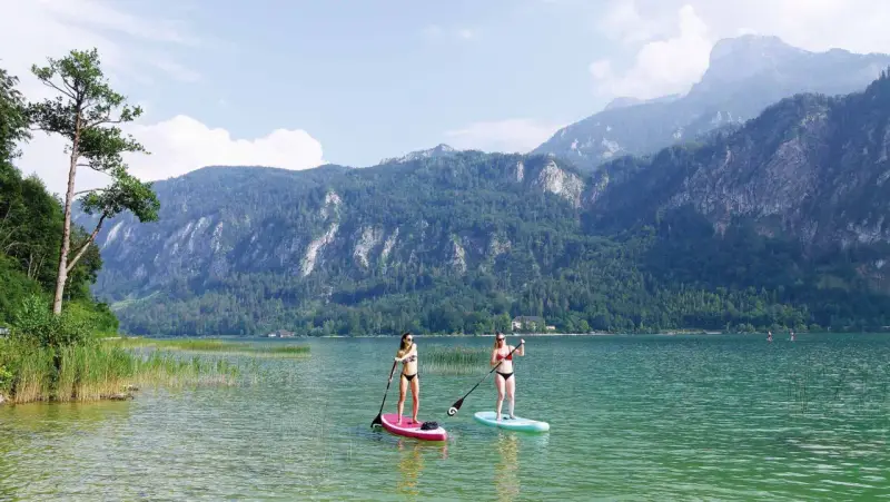 Zwei Frauen genießen Stand-Up-Paddling auf einem ruhigen See vor einer Bergkulisse. Perfekte sup spots für einen entspannten Tag.