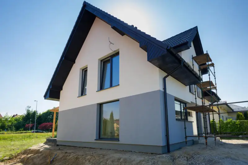 Nowy dom z dwukolorową elewacją, biało-szarą, to świetny pomysł na elewację domu. Widać rusztowania, co sugeruje trwające prace.