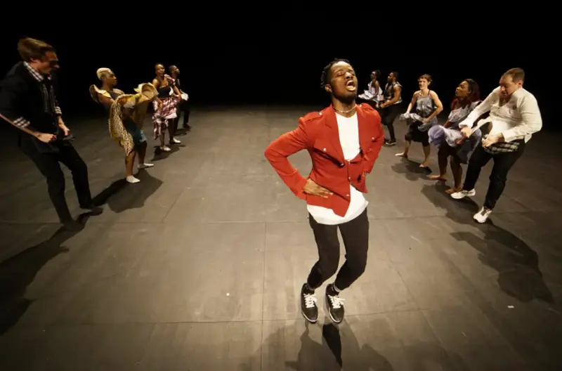 Un danseur afro 93, en veste rouge, mène une troupe sur scène. Les danseurs, aux tenues variées, exécutent des mouvements synchronisés.