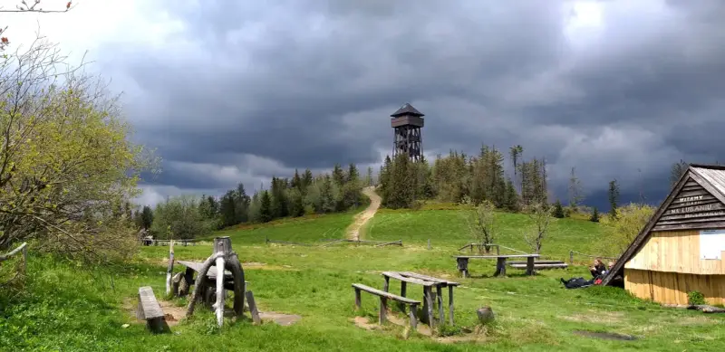 Drewniana wieża widokowa na Lubaniu. Trasa prowadzi przez zielone wzgórza pod burzowym niebem.