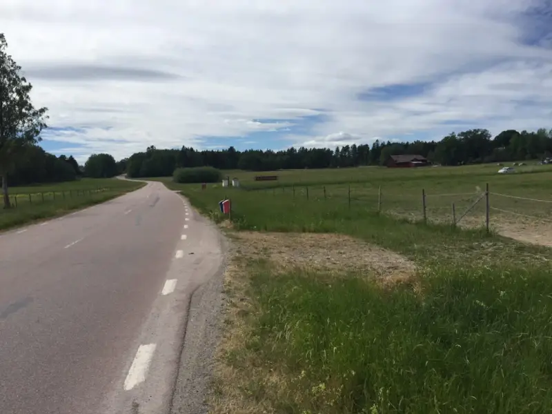 Vacker väg kantad av grönska, perfekt för att utforska Sveriges cykelleder. En röd lada skymtar i bakgrunden.