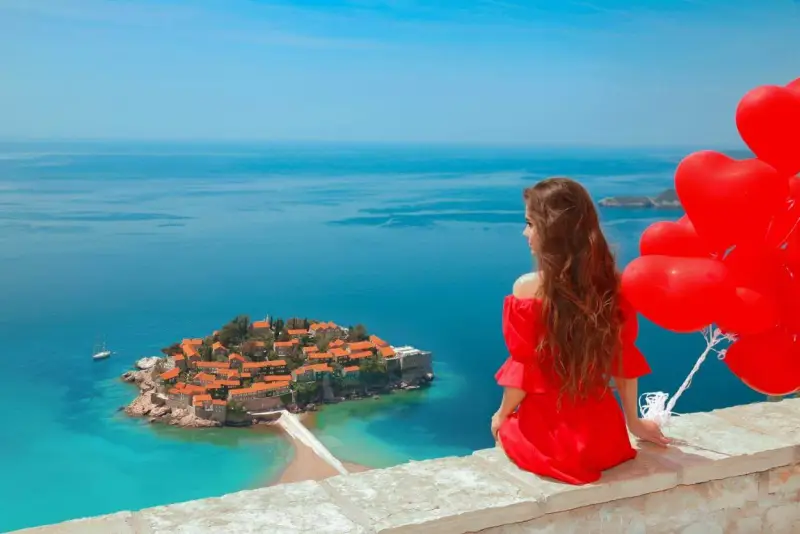 Marzenie o czarnogóra all inclusive samolotem? Kobieta w czerwonej sukni z sercami w ręku podziwia wyspę Sveti Stefan.