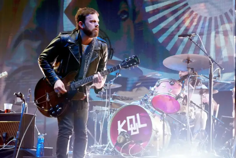Gitarzysta Kings of Leon Polska w skórzanej kurtce gra na scenie. W tle perkusja z logo KQL.