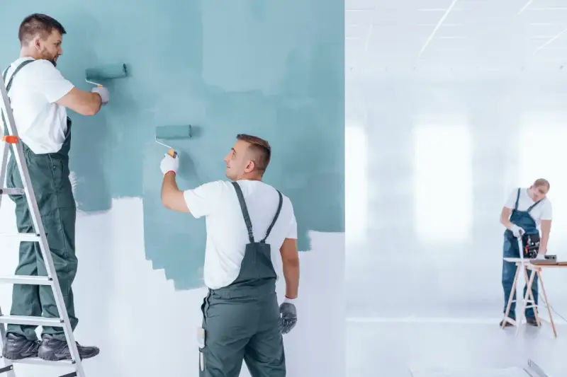 Malowanie ścian na miętowo. Dwóch fachowców na drabinie i stojącego maluje, trzeci pracuje przy stole. Sprawdź, ile za metr malowania ścian!