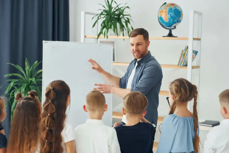 Nauczyciel tłumaczy coś dzieciom przy tablicy. To mogą być dni dyrektorskie, podczas których uczniowie poznają szkołę.