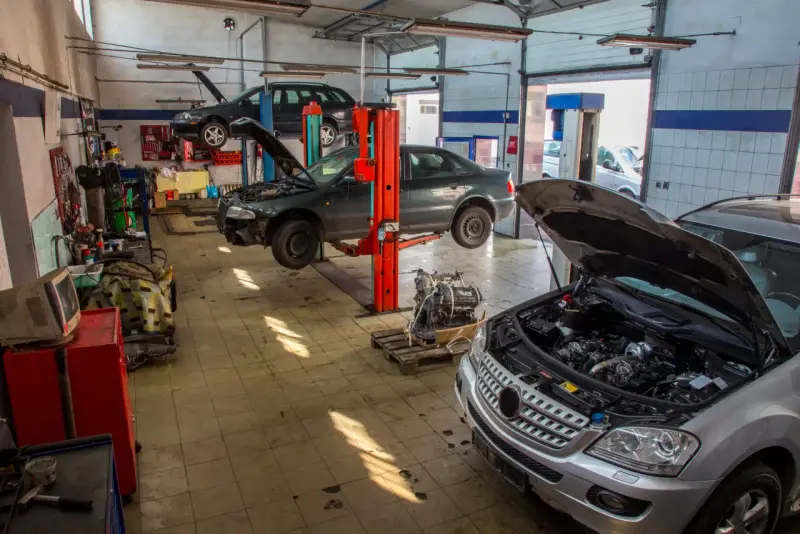 Warsztat samochodowy z kilkoma autami na podnośnikach, w tym ciemnym sedanem, który wygląda jak p1351 peugeot. Silnik wyjęty z jednego z pojazdów.