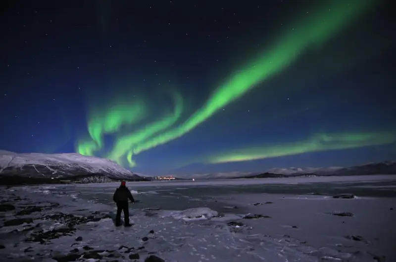 En ensam person står på en isig strand och ser på det magiska abisko norrsken som dansar över himlen.