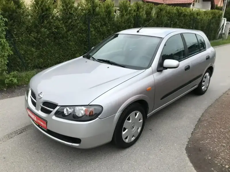 Srebrny Nissan Almera hatchback stoi na asfaltowej drodze, otoczony zielenią.