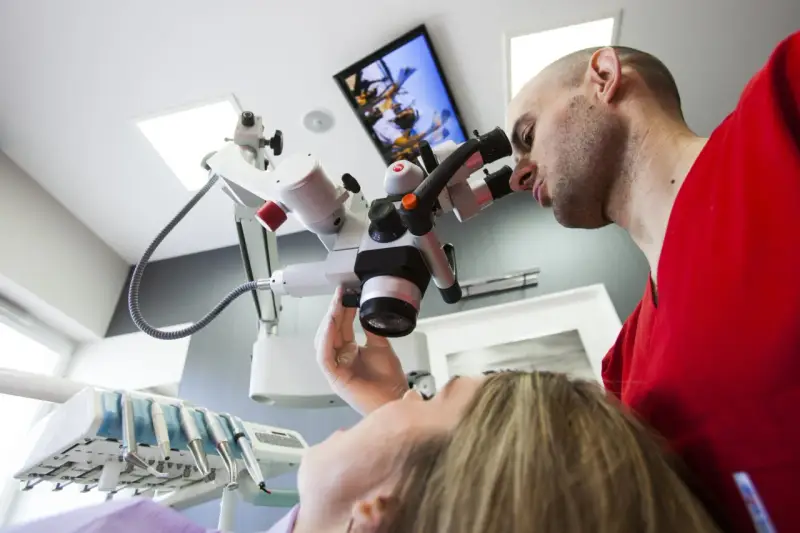 Dentysta w czerwonej bluzie bada pacjentkę przez mikroskop stomatologiczny.
