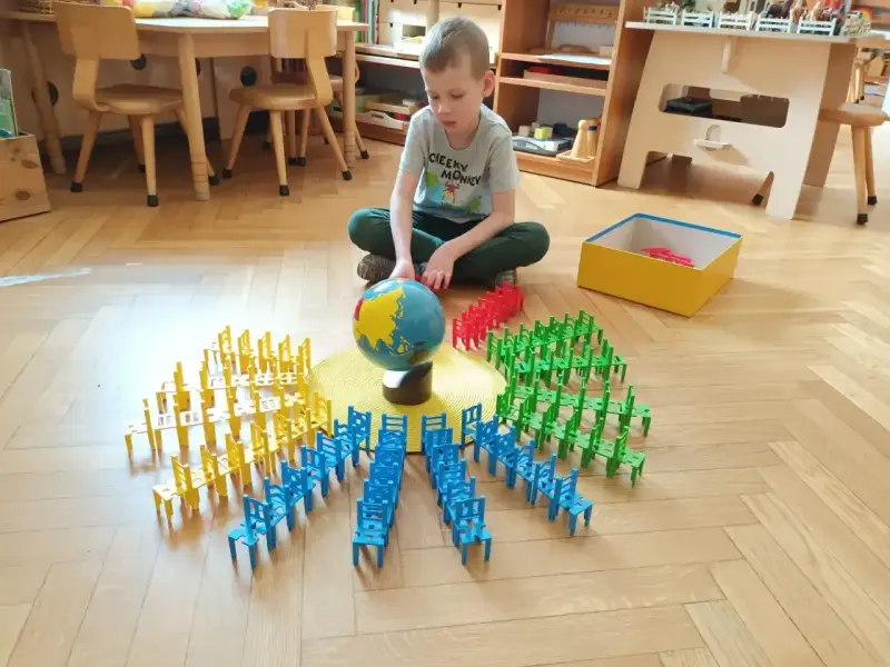 Chłopiec bawi się w przedszkolu Montessori, układając kolorowe krzesełka wokół globusa. To pokazuje, montessori co to za metoda nauczania.