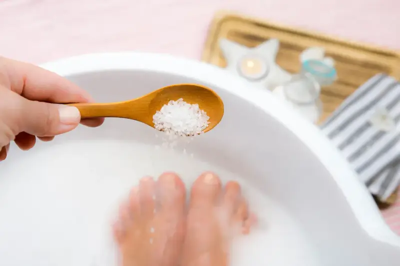 A hand adds epsom salt and baking soda to a foot soak, with feet submerged in the milky water.
