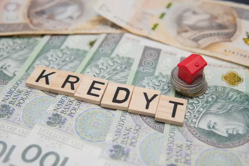 Litery układające słowo "KREDYT" na banknotach. Mały czerwony domek na stosie monet symbolizuje pomoc w spłacie kredytu, np. z funduszu wsparcia kredytobiorców.