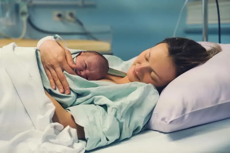 Eine Mutter hält ihr Neugeborenes im Arm, glücklich und erschöpft. Die Szene zeigt, wie lange nach Geburt im Krankenhaus die erste Bindung entsteht.