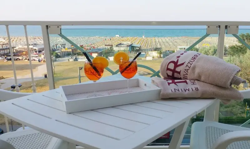 Due aperitivi arancioni e asciugamani con logo "Hotel Rivazzurra Rimini" sul tavolo, con vista sul rivazzurra mare e spiaggia attrezzata.