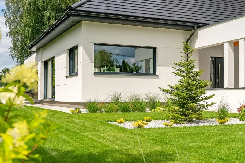 Nowoczesny dom z białym tynkiem i dużymi oknami PCV. Piękny ogród z trawnikiem i iglakami.