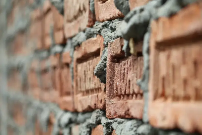 Zbliżenie na ceglaną ścianę w budowie. Widać nierównomiernie nałożoną zaprawę i teksturę cegieł. Zastanawiasz się, ile łopat piasku na worek cementu?