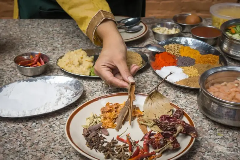 Ręka wybiera cynamon z talerza przypraw, przygotowując autentyczną kuchnię nepalską. Widać chili, mąkę, pastę imbirową i kolorowe przyprawy.