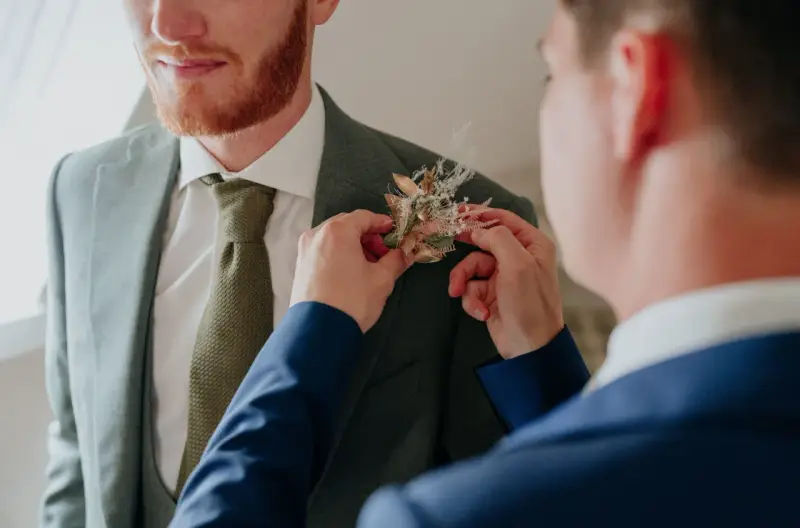 Un homme ajuste une boutonnière sur le costume d'un autre. Divers types de boutonnières existent, celle-ci est composée de fleurs séchées et de feuillages dorés.