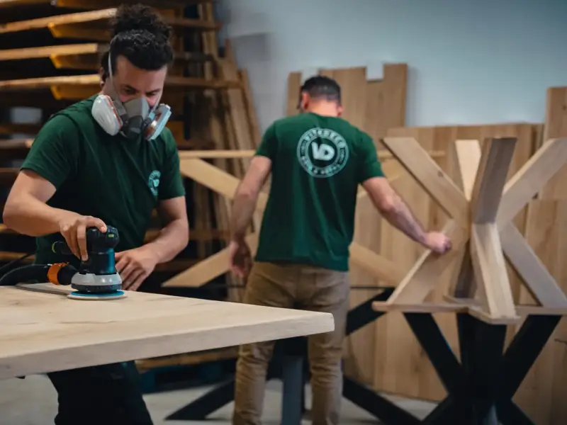 Un artisan, masqué, utilise une ponceuse pour poncer une table en bois. Un autre artisan travaille sur un pied de table en arrière-plan.