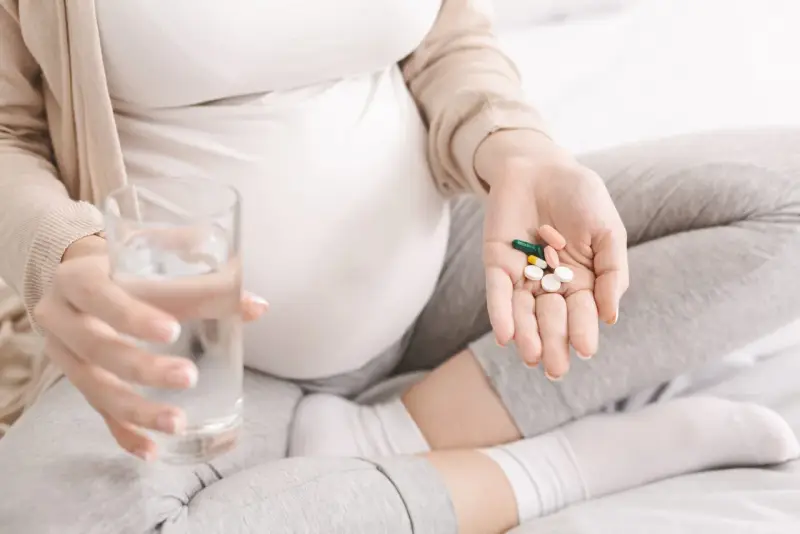 Ciężarna kobieta trzyma garść tabletek i szklankę wody, szukając odpowiedzi na pytanie: co na katar w ciąży.
