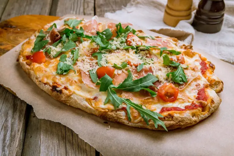 Pinsa co to? Aromatyczna, podłużna pizza z rukolą, pomidorkami i szynką parmeńską, posypana parmezanem.