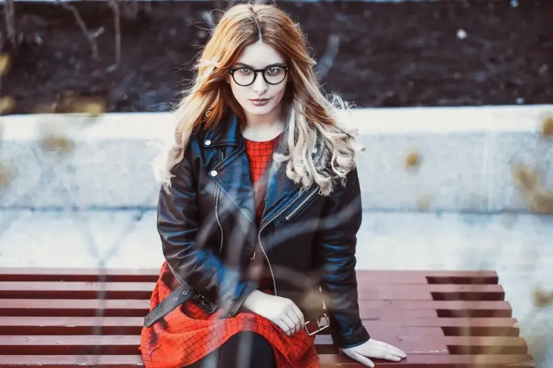Dziewczyna w okularach i czerwonej sukience siedzi na ławce, ubrana w stylową kurtkę Dolce & Gabbana.