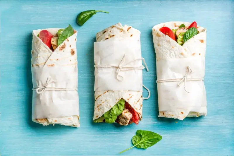 Trzy keto tortille z siemienia lnianego, zawinięte w papier i przewiązane sznurkiem, z widocznym nadzieniem z kurczaka, pomidorów i szpinaku.