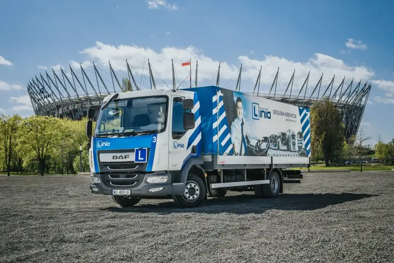 Ciężarówka szkoleniowa z literą "L" na masce, gotowa do nauki jazdy kat. C. W tle stadion.