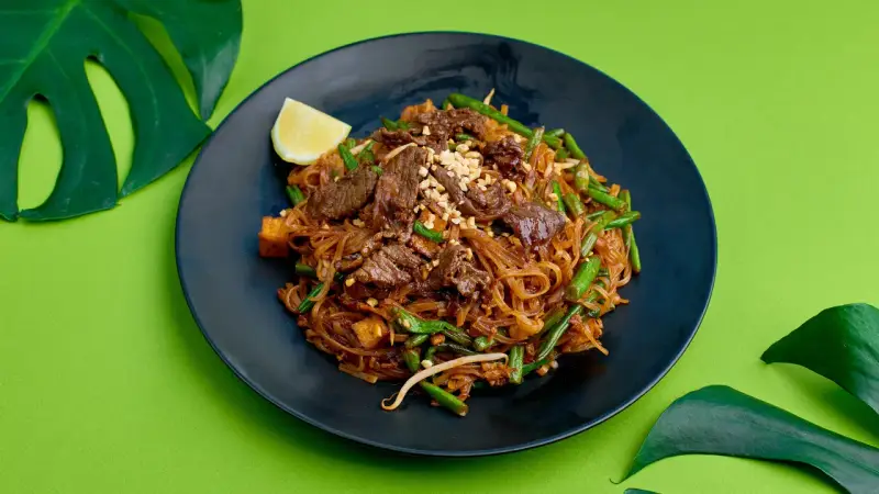 Pyszne pad thai z wołowiną, fasolką szparagową i orzeszkami na czarnym talerzu, ozdobione ćwiartką cytryny i liśćmi monstery.
