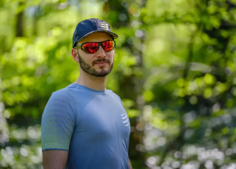 Mężczyzna w czapce i sportowych okularach przeciwsłonecznych, z odbiciem lasu w soczewkach.