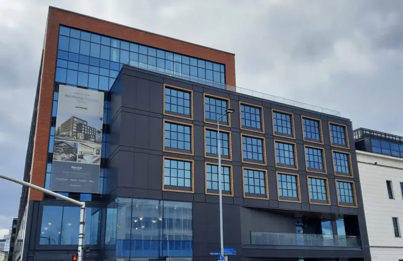 Nowoczesny budynek z cegły i ciemnej fasady, z banerem zapowiadającym otwarcie Occidental Powisle Hotel, części sieci Barceló.