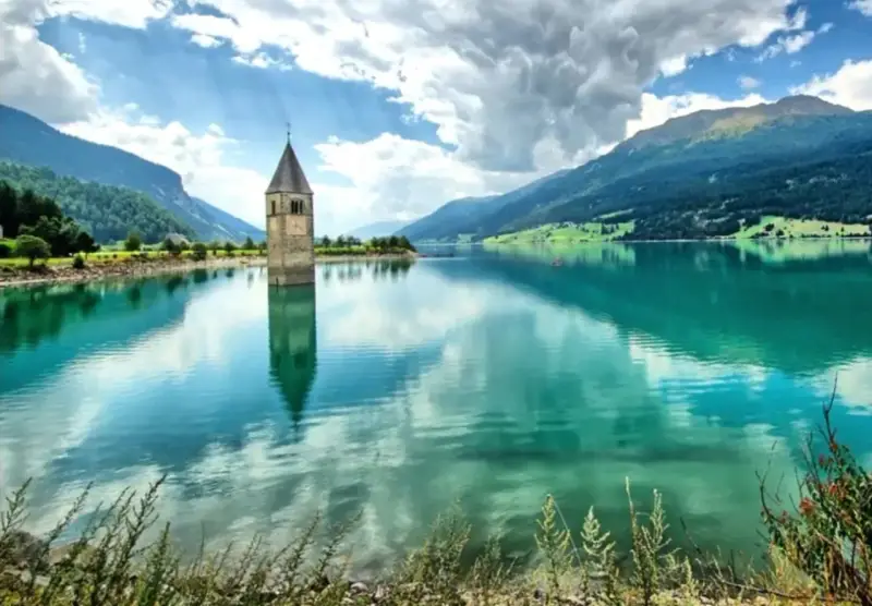 Il lago del campanile, con la sua torre sommersa, riflette montagne verdi e un cielo nuvoloso.