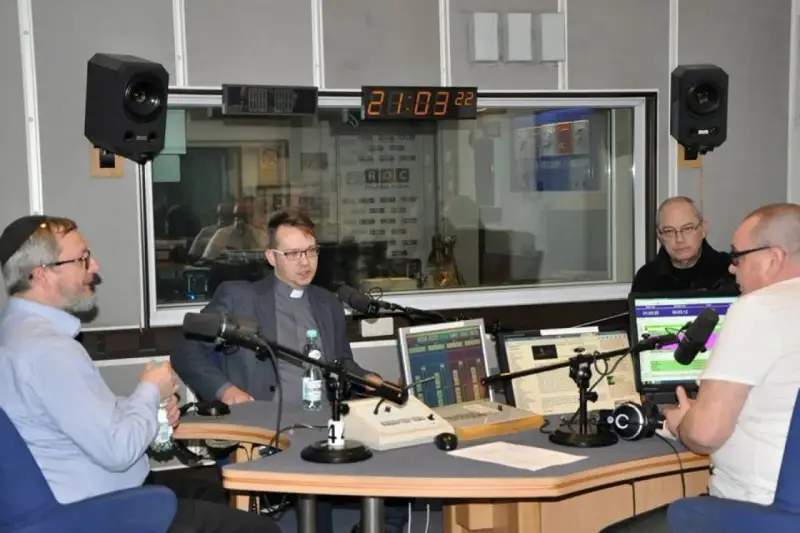 W studiu radiowym czterech mężczyzn, w tym ksiądz i osoba w jarmułce, rozmawia przy mikrofonach. W tle widać zegar i logo RDC.
