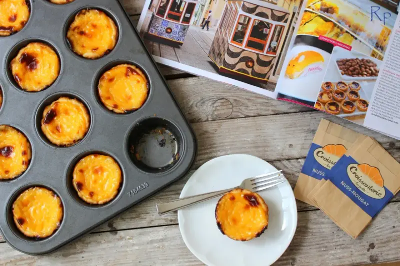 Pyszne, lekko przypieczone pastel de nata w blaszce i na talerzyku. Obok magazyn z widokiem na Lizbonę i tramwaje.