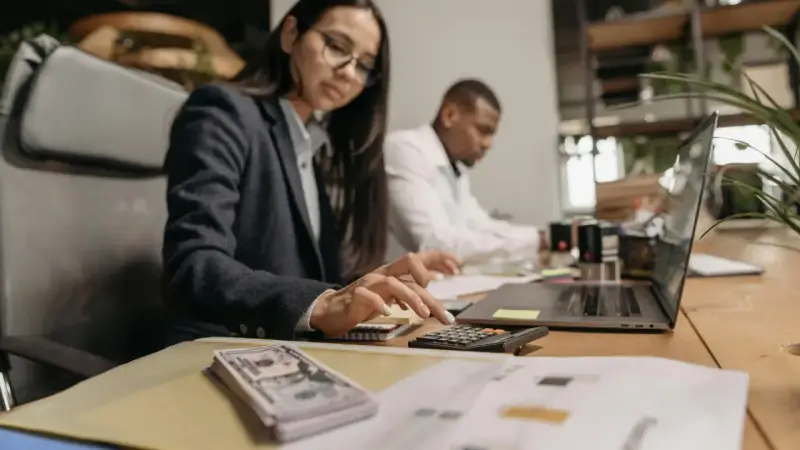 Kobieta w okularach liczy na kalkulatorze, obok stos banknotów i laptop.