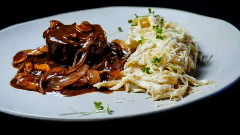Mięso w sosie z cebulą i grzybami, przypominającymi podgrzybki, podane z kremowym makaronem posypanym parmezanem i kiełkami.