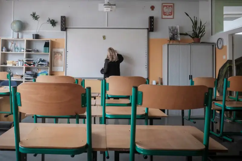 Nauczycielka przy tablicy, zastanawia się, ile zarabia przedszkolanka. Puste ławki w sali lekcyjnej.