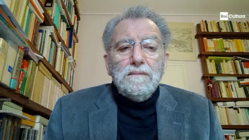 Uomo anziano con barba grigia, occhiali e giacca, circondato da scaffali pieni di libri. Il suo sguardo intenso suggerisce che ei fu significato.