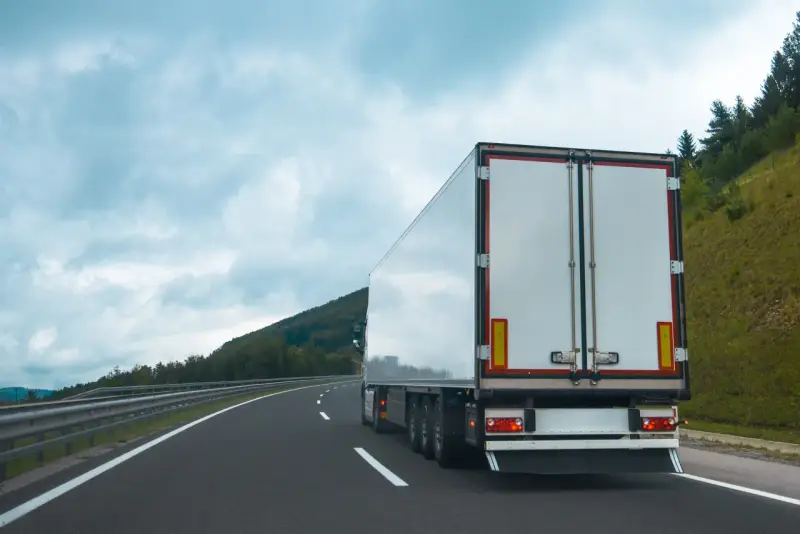 Ciężarówka na autostradzie, z tyłu widoczne białe drzwi naczepy. W tle góry i chmury. Tablice wyróżniające przepisy są częścią oznakowania pojazdu.