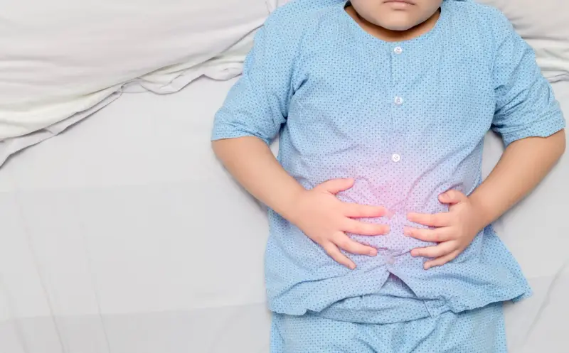 Niño con pijama azul, acostado en cama blanca, se agarra la barriga con las manos. Se visualiza un área rosada en su abdomen, indicando dolor barriga niños.