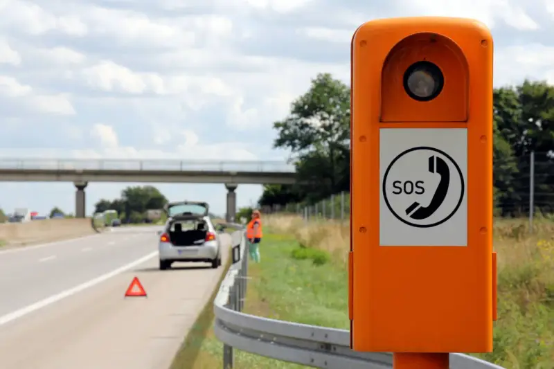 Ihr Fahrzeug ist auf der Autobahn liegen geblieben. Eine Frau in Warnweste steht neben dem offenen Kofferraum, ein Warndreieck liegt auf der Fahrbahn. Eine Notrufsäule steht im Vordergrund.