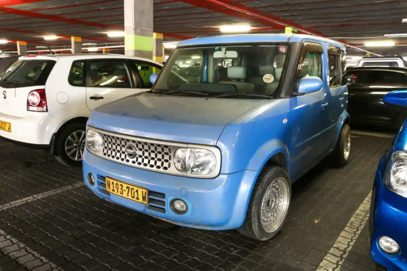 Niebieski Nissan Cube z charakterystycznym grillem i tablicą rejestracyjną N193-701 W, zaparkowany obok białego Volkswagena.