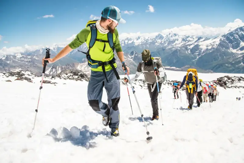 Un gruppo di escursionisti sta camminando sulla neve in montagna, con bastoncini da trekking e zaini, sotto un cielo azzurro.