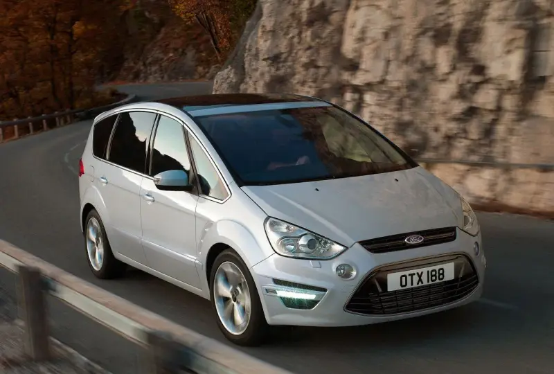 Biały Ford S-Max 2.0 TDCI pokonuje zakręt na malowniczej drodze wśród jesiennych drzew i skał.