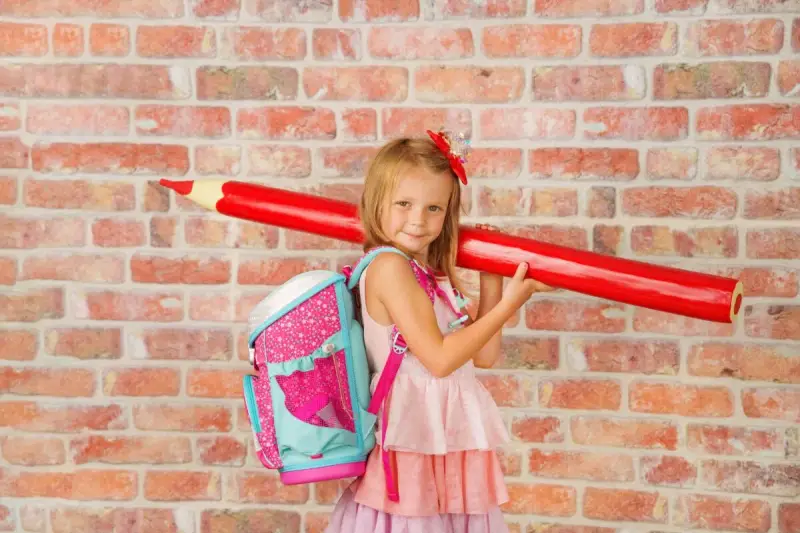 Ein Mädchen mit pinkem Rucksack und riesigem roten Stift vor Ziegelwand. Bereit für die Einschulung Berlin 2025!
