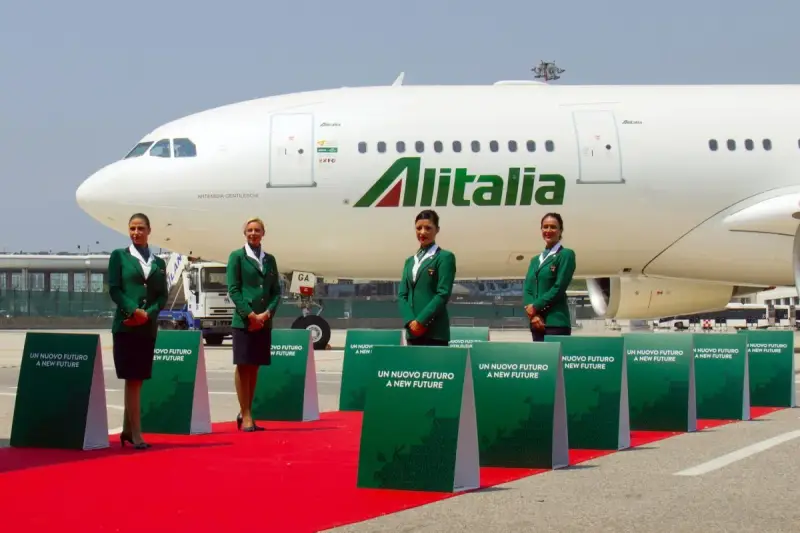 Cztery stewardessy w zielonych uniformach stoją na czerwonym dywanie przed samolotem Alitalia.