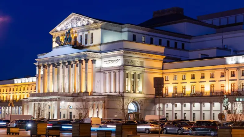 Teatr Wielki w Warszawie, najstarszy teatr w Polsce, nocą. Monumentalna fasada z kolumnami i rzeźbami, oświetlona ciepłym światłem, z samochodami w ruchu ulicznym przed budynkiem.